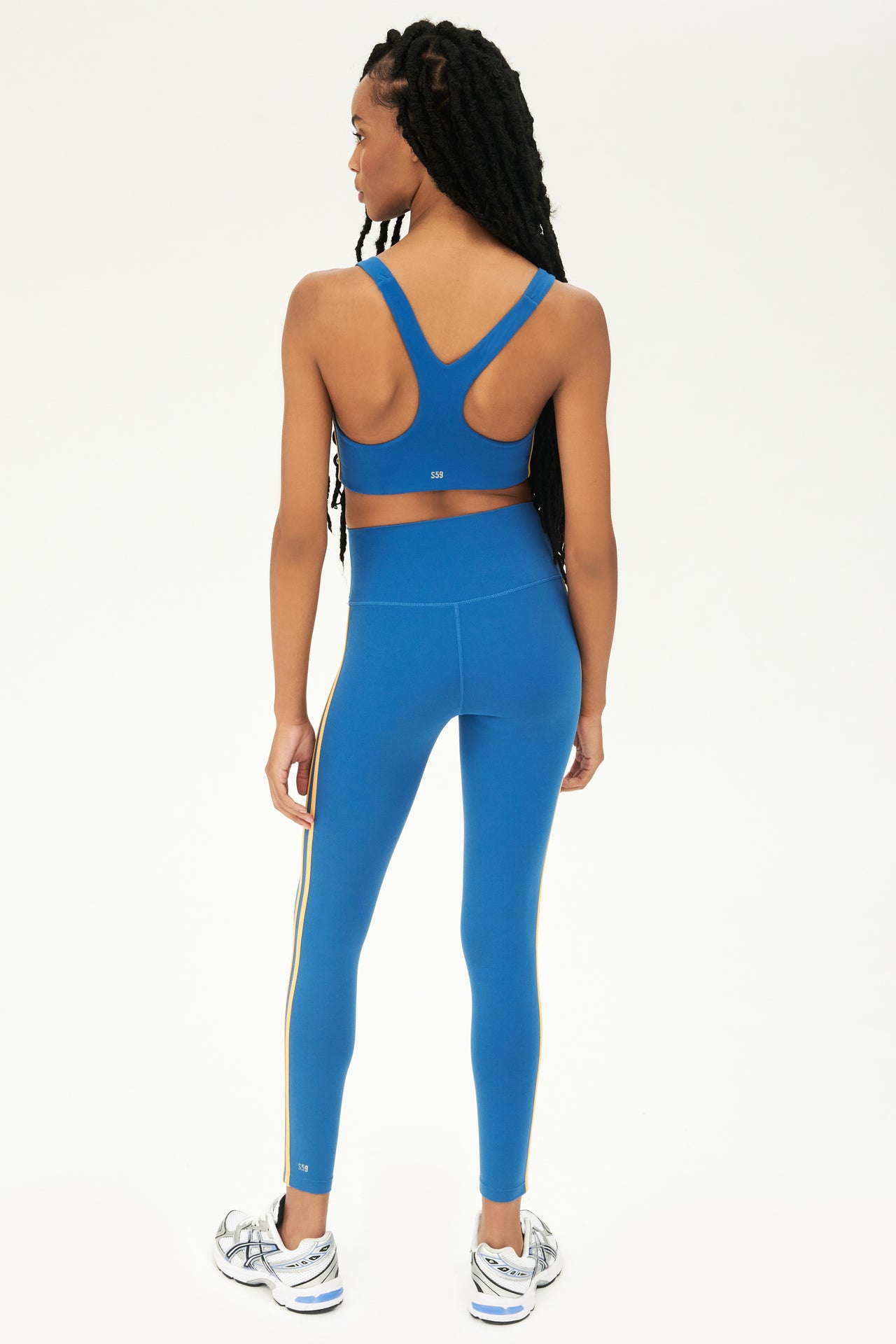Full back view of girl wearing blue sports bra with two thin yellow stripes down the side and blue leggings with white shoes