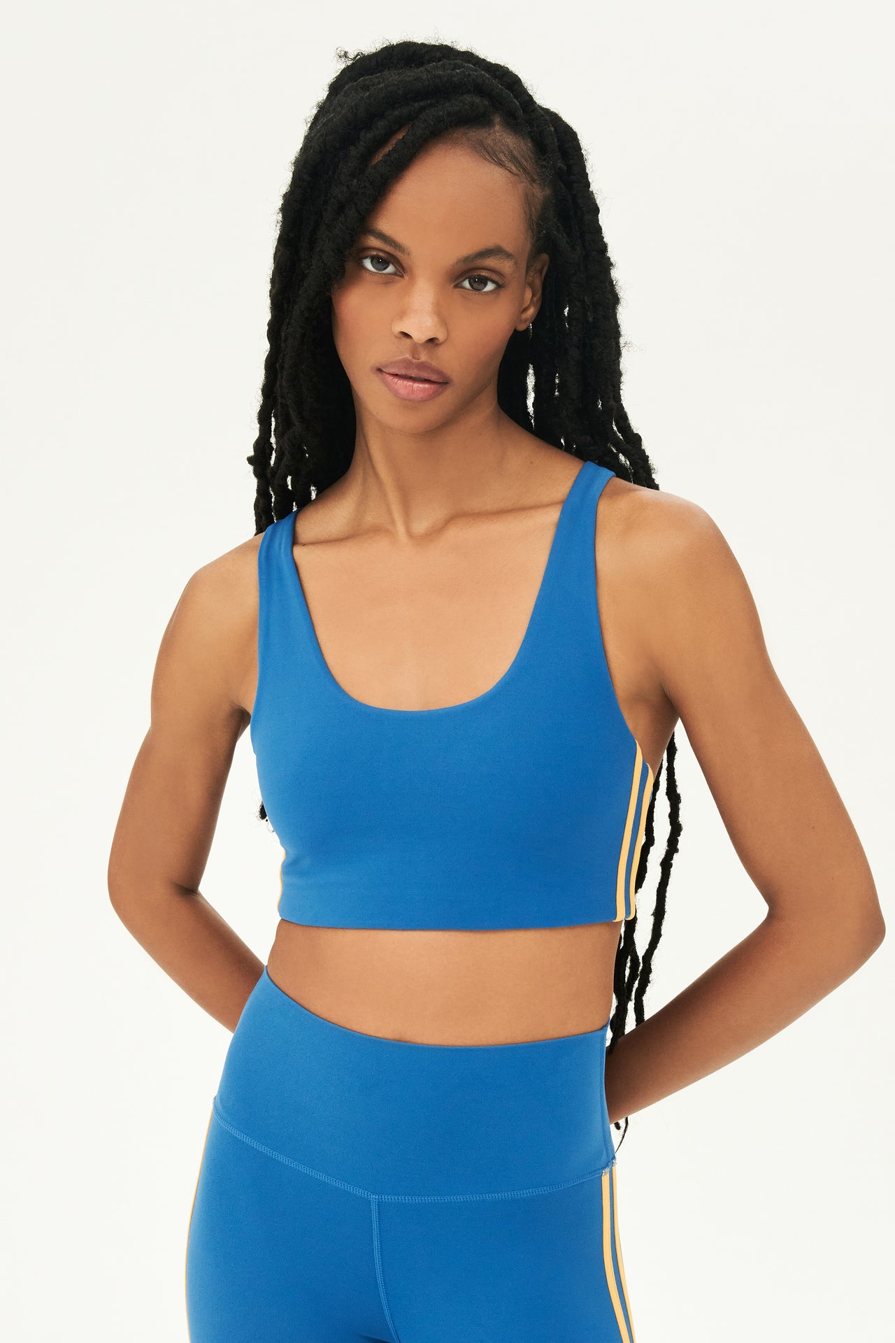 Front view of girl wearing blue sports bra with two thin yellow stripes down the side and blue leggings 