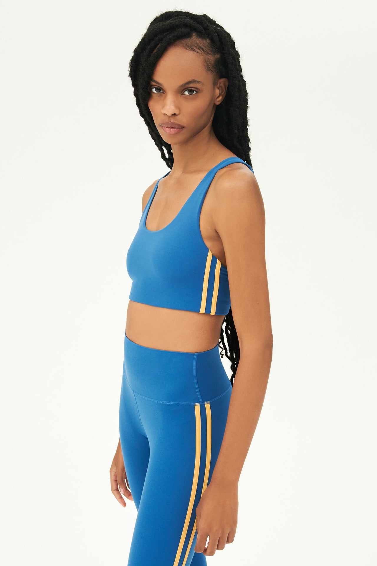 Side view of girl wearing blue sports bra with two thin yellow stripes down the side and blue leggings 