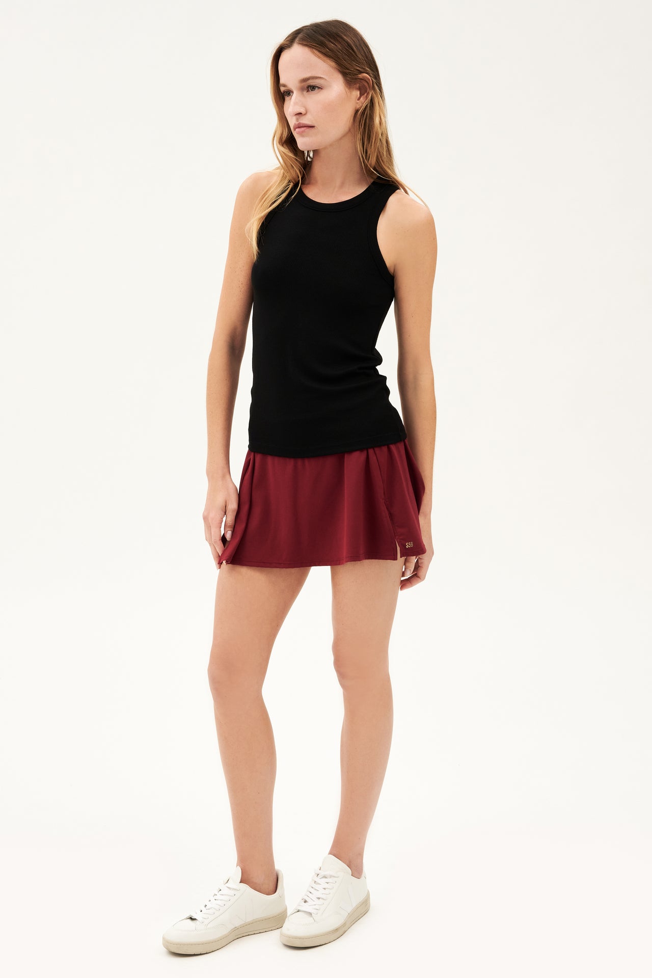 Full front side view of woman with dark blonde hair wearing a blank tank top and dark red skort with white shoes 