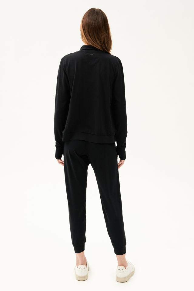 The back view of a woman wearing a SPLITS59 Rain Airweight Jacket - Black and joggers, ready for a Pilates class.