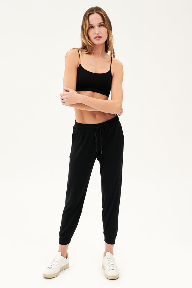 Full front view of girl wearing black sweatpants with black tie around waistband and a black sports bra with white shoes 