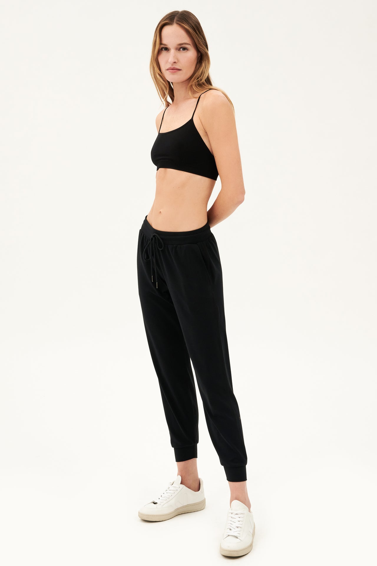Full side view of girl wearing black sweatpants with black tie around waistband and a black sports bra with white shoes 