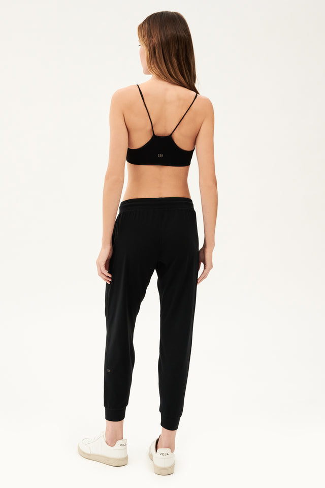 Full back view of girl wearing black sweatpants with black tie around waistband and a black sports bra with white shoes 