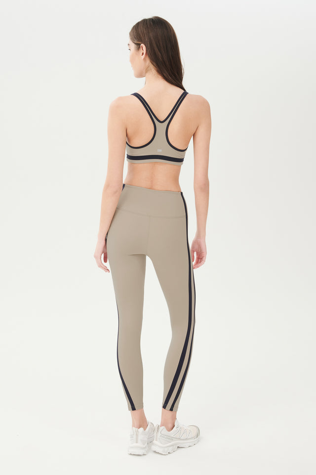 Full back view of girl wearing dark light brown sports bra with black colored stripe along the ribs and black hem around the arms and neck with dark light brown leggings with two black stripes down the side and white shoes