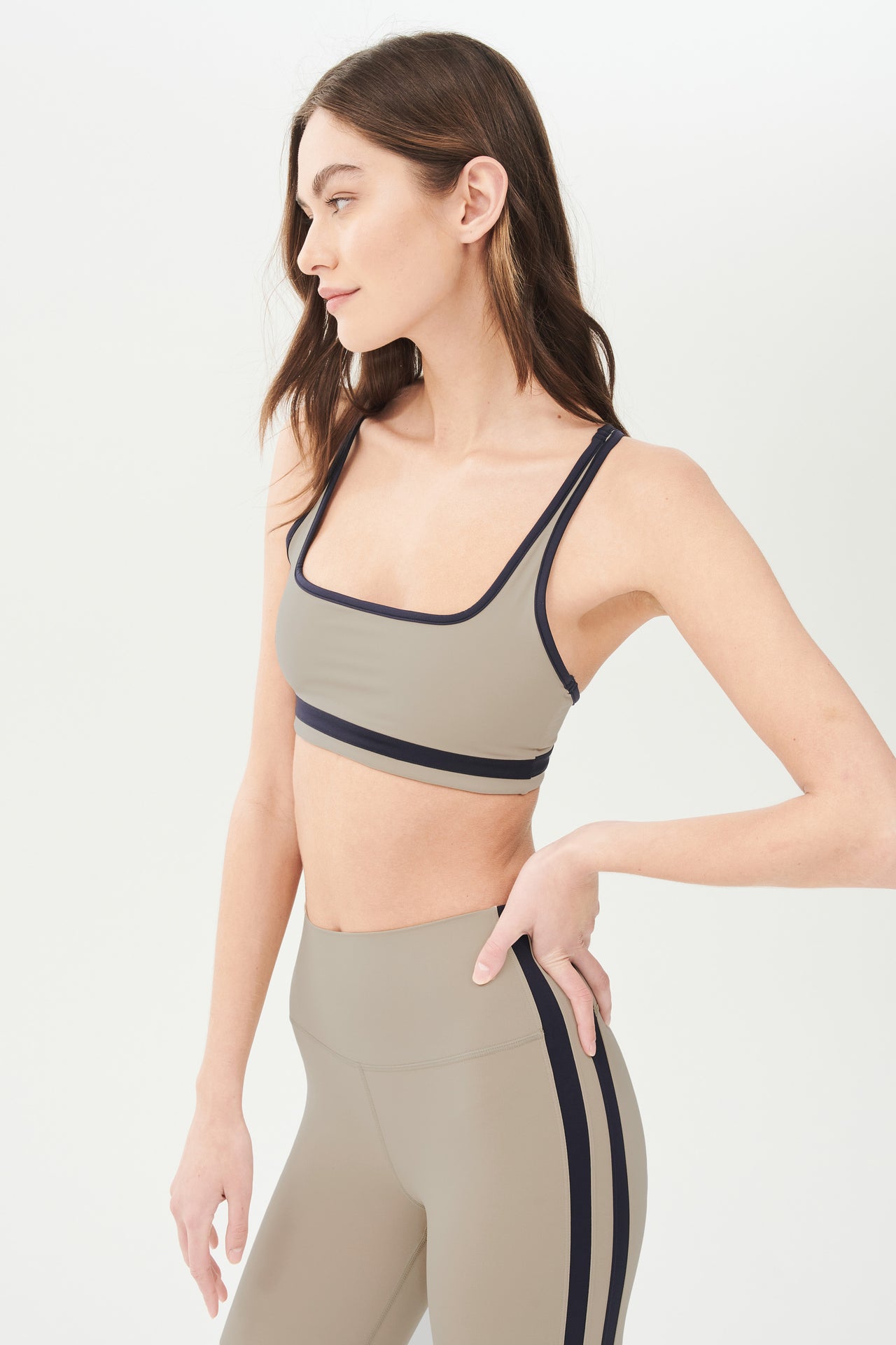 Side view of girl wearing dark light brown sports bra with black colored stripe along the ribs and black hem around the arms and neck with dark light brown leggings with two black stripes down the side 