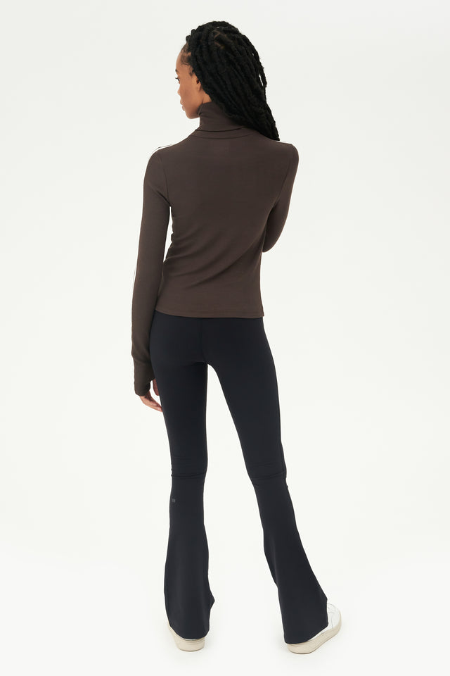The back view of a woman wearing black leggings and a SPLITS59 Jackson Rib Turtleneck in Dark Chocolate/Creme, ideal for a Pilates session.