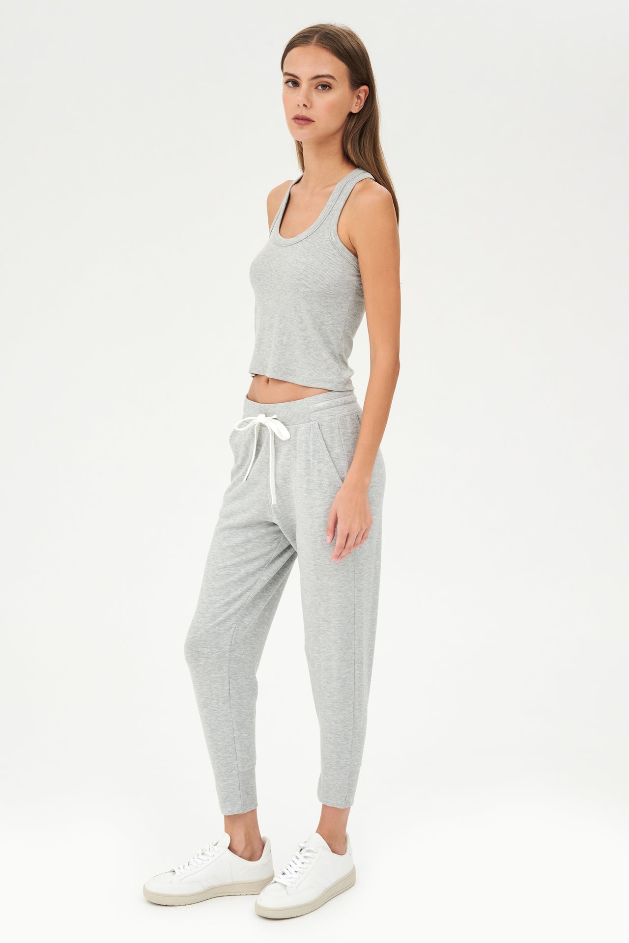 Full side view of girl wearing a ribbed grey cropped tank top and grey sweatpants with a white tie along the waistband with white shoes