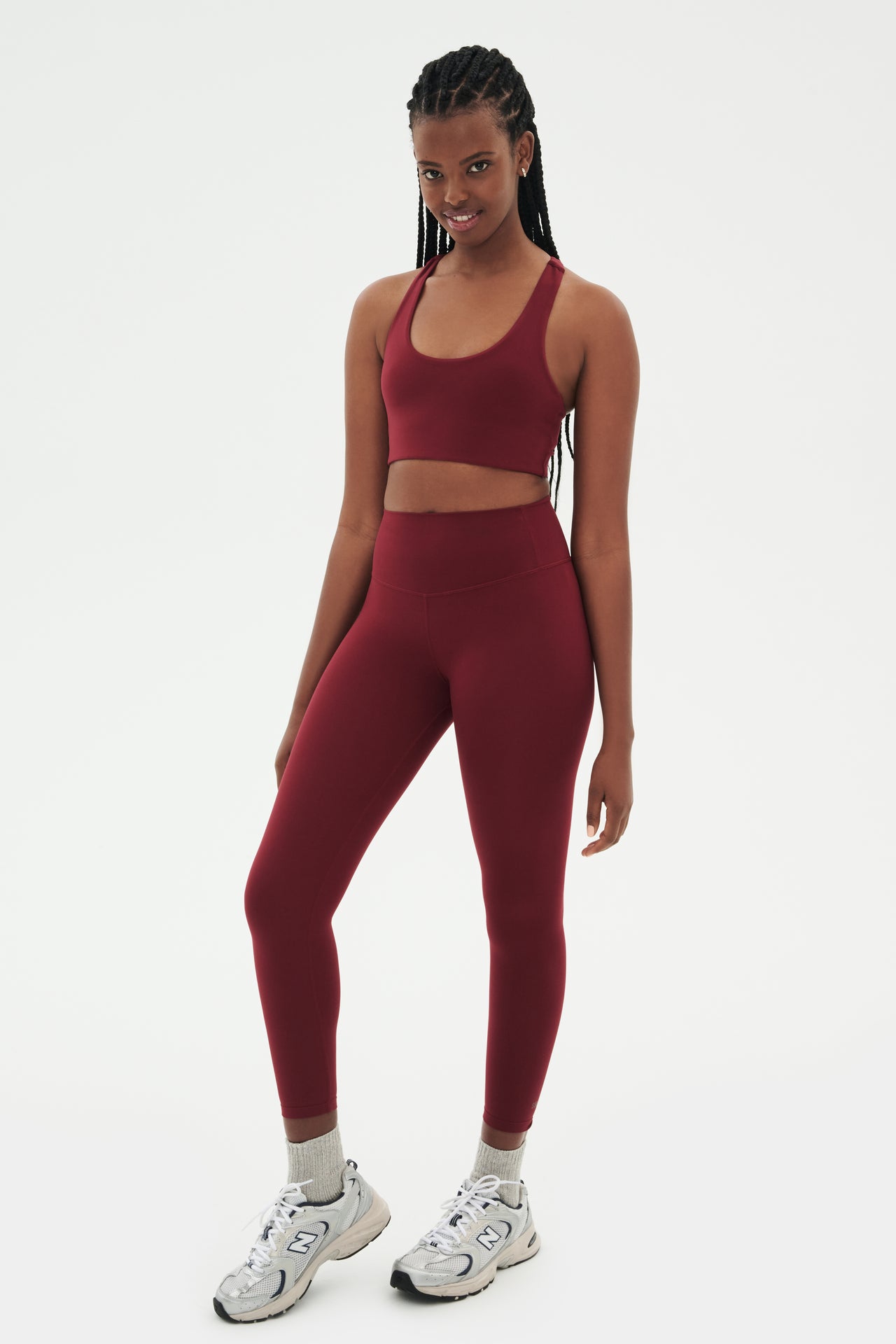 Full side view of girl wearing highwasited dark red leggings with dark red sports bra and grey shoes