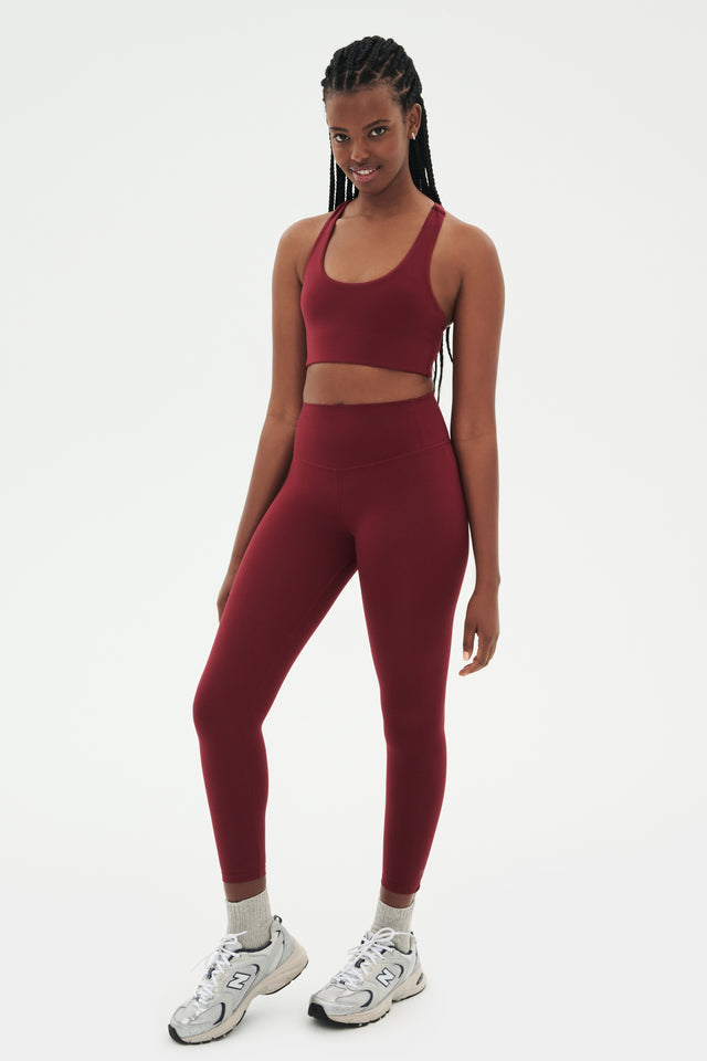 Full side view of girl wearing highwasited dark red leggings with dark red sports bra and grey shoes