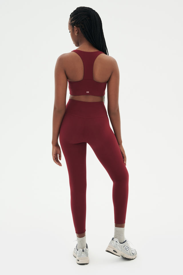 The back view of a woman wearing a burgundy SPLITS59 Sara Airweight Bra - Bordeaux and leggings for hot yoga.