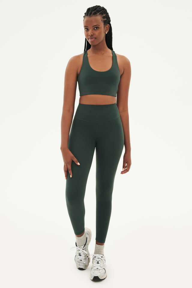 Full front view of woman with black braids wearing dark green leggings and dark green bra paired with white shoes with black stripes