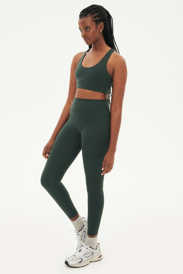 Full front side view of woman with black braids wearing dark green leggings and dark green bra  paired with white shoes with black stripes