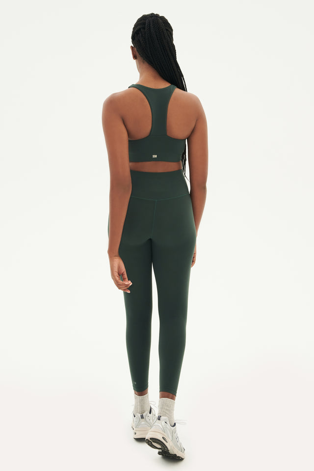 Full back view of woman with black braids wearing dark green leggings and dark green bra with racerback paired with white shoes with black stripes