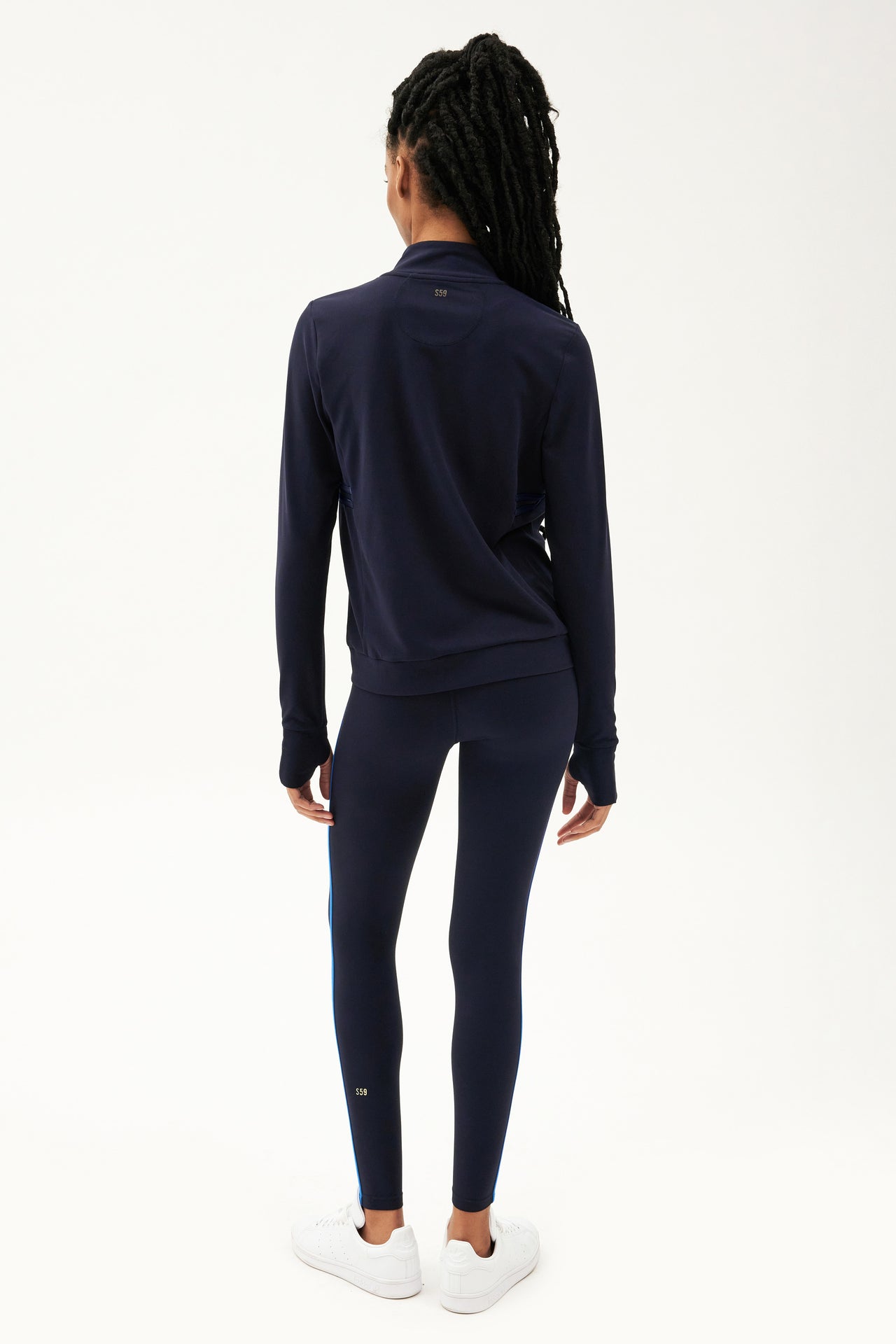 The back view of a woman wearing a SPLITS59 Rain Airweight Jacket in Indigo and sleek leggings designed for hot yoga.