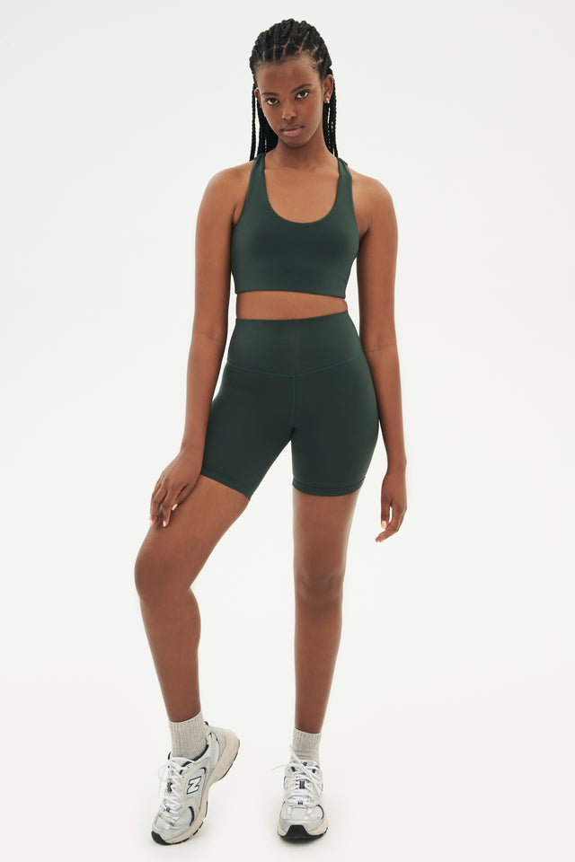 Full front view of girl wearing highwaisted mid thigh dark dark green bike shorts with dark green sports bra and white shoes