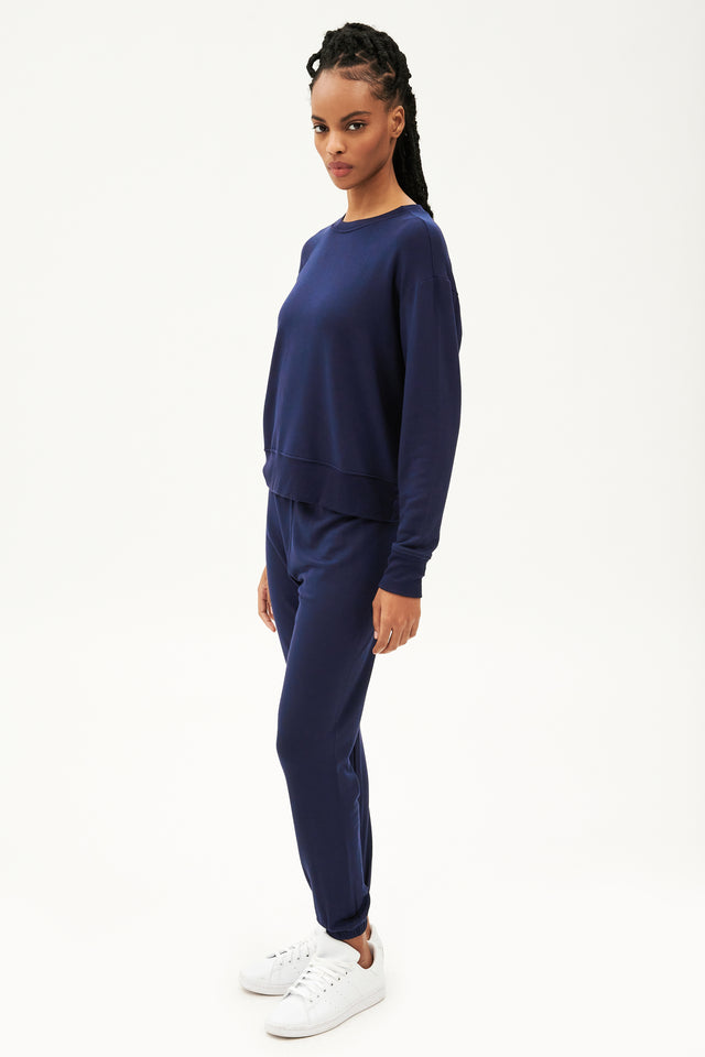 Full front side view of woman with black braids wearing a dark blue crewneck sweatshirt  with dark blue joggers paired with white shoes 