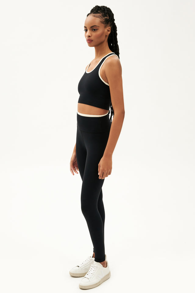 Full side view of girl wearing black leggings with thin white band around waist and black tank top with white shoes
