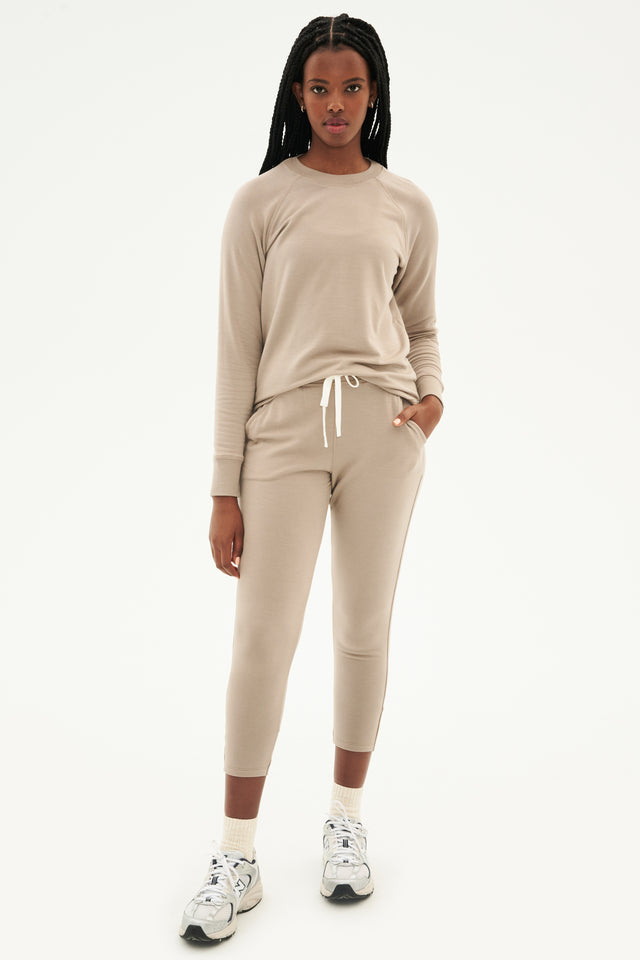 Front full view of woman with black braids wearing light brown creme tone crewneck sweatshirt and sweatpant with tapered leg and above ankle length with white drawstring and side hip pockets paired with white shoes with black stripes