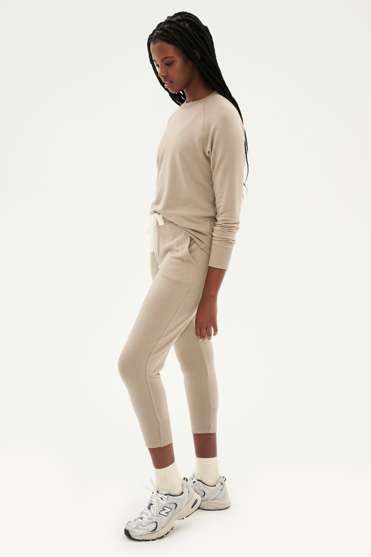 Front side view of woman with black braids wearing light brown creme tone crewneck sweatshirt and sweatpant with tapered leg and above ankle length with white drawstring paired with white shoes with black stripes