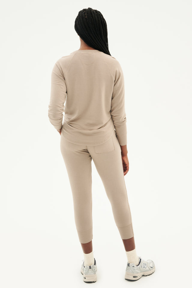 Full back view of woman with black braids wearing light brown creme tone crewneck sweatshirt and sweatpant with tapered leg and above ankle length with back pocket paired with white shoes with black stripes