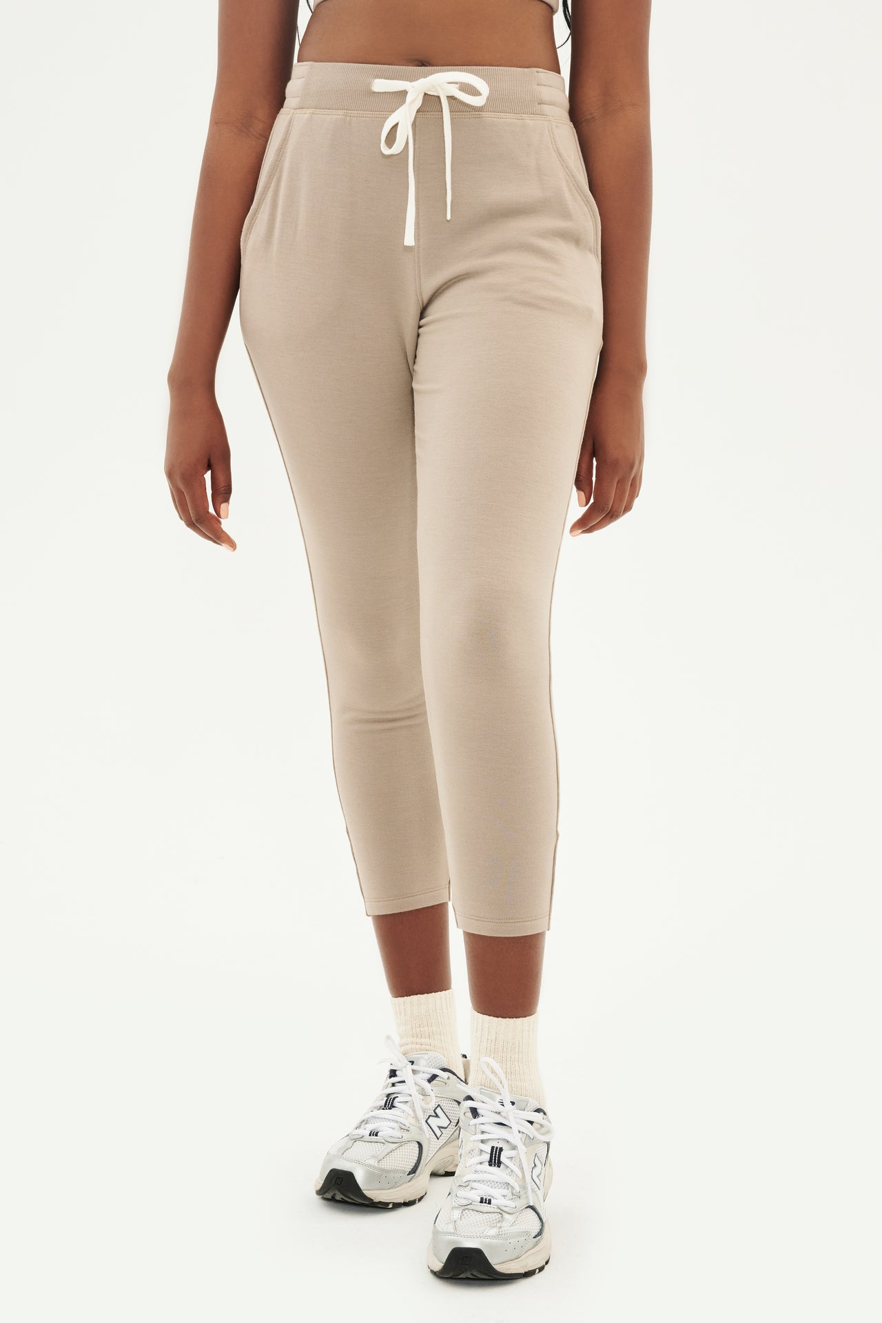 Front view of woman wearing light brown creme tone crewneck sweatshirt and sweatpant with tapered leg and above ankle length with white drawstring and side hip pockets paired with white shoes with black stripes