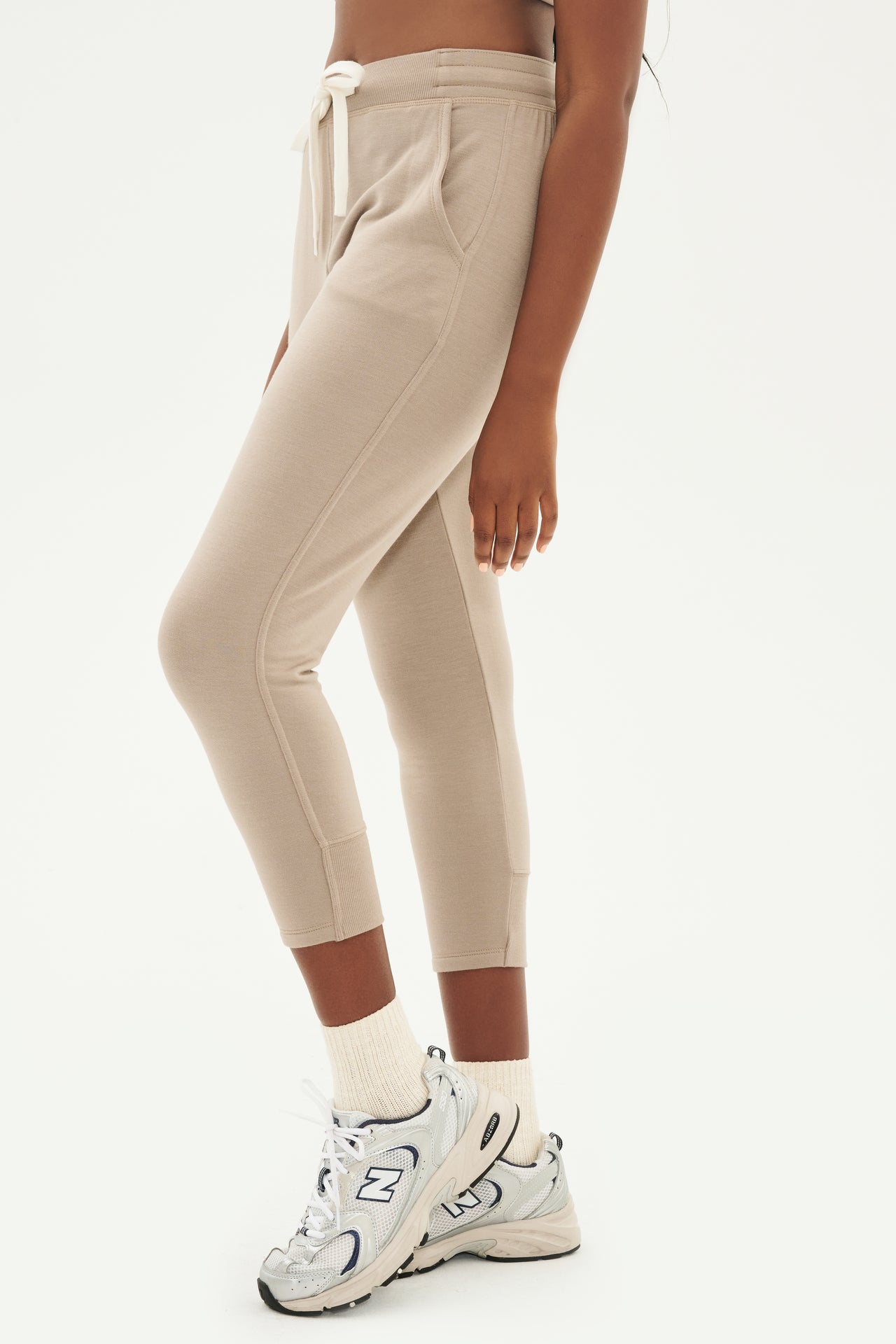 Front side view of woman wearing light brown creme tone crewneck sweatshirt and sweatpant with tapered leg and above ankle length with white drawstring and side hip pockets paired with white shoes with black stripes