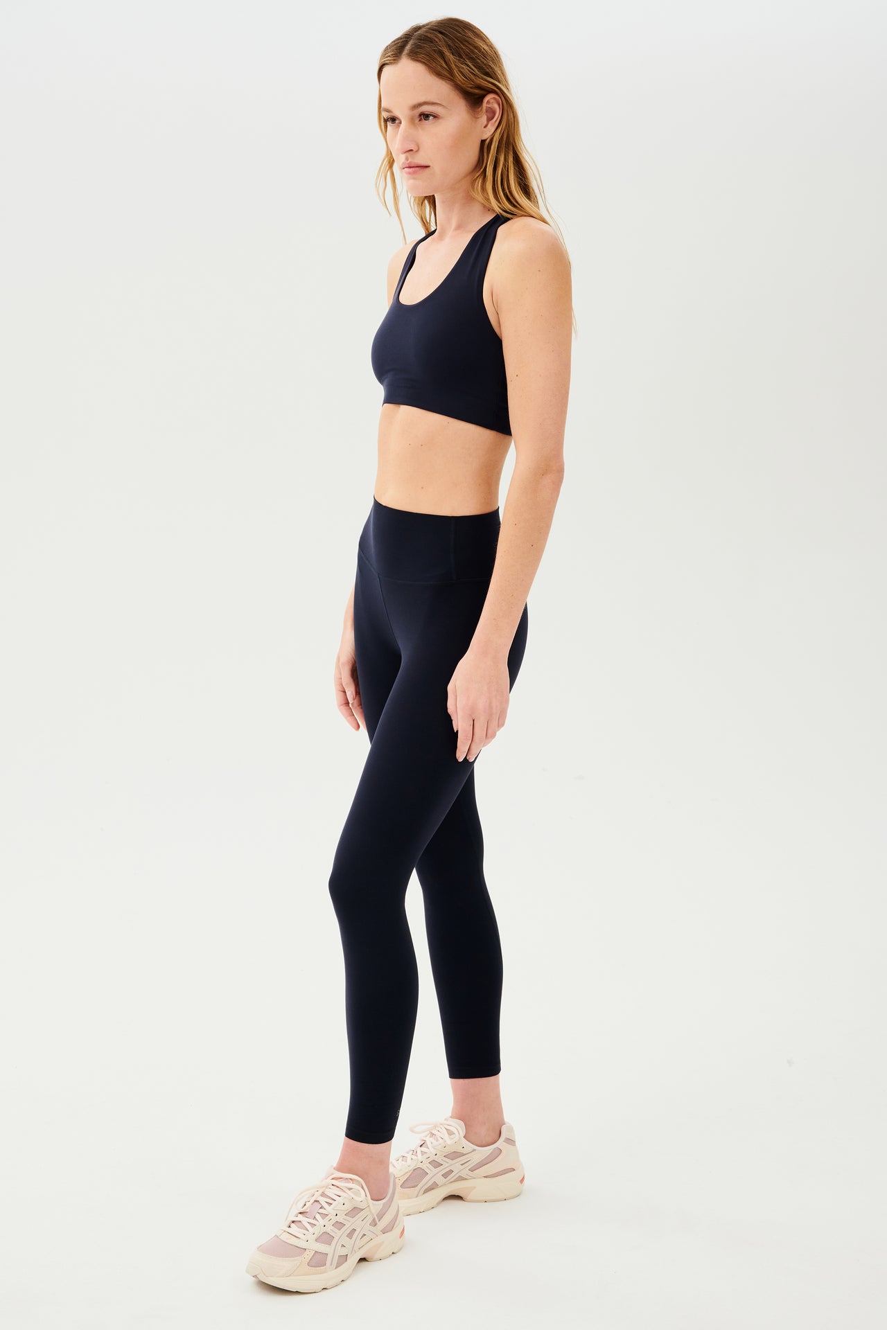 Full side view of girl wearing highwasited dark blue leggings with dark blue sports bra and pink and cream shoes