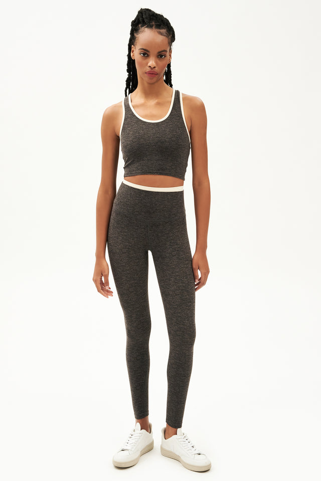 Full front view of girl wearing dark grey leggings with thin white band around waist and dark grey tank top with white shoes