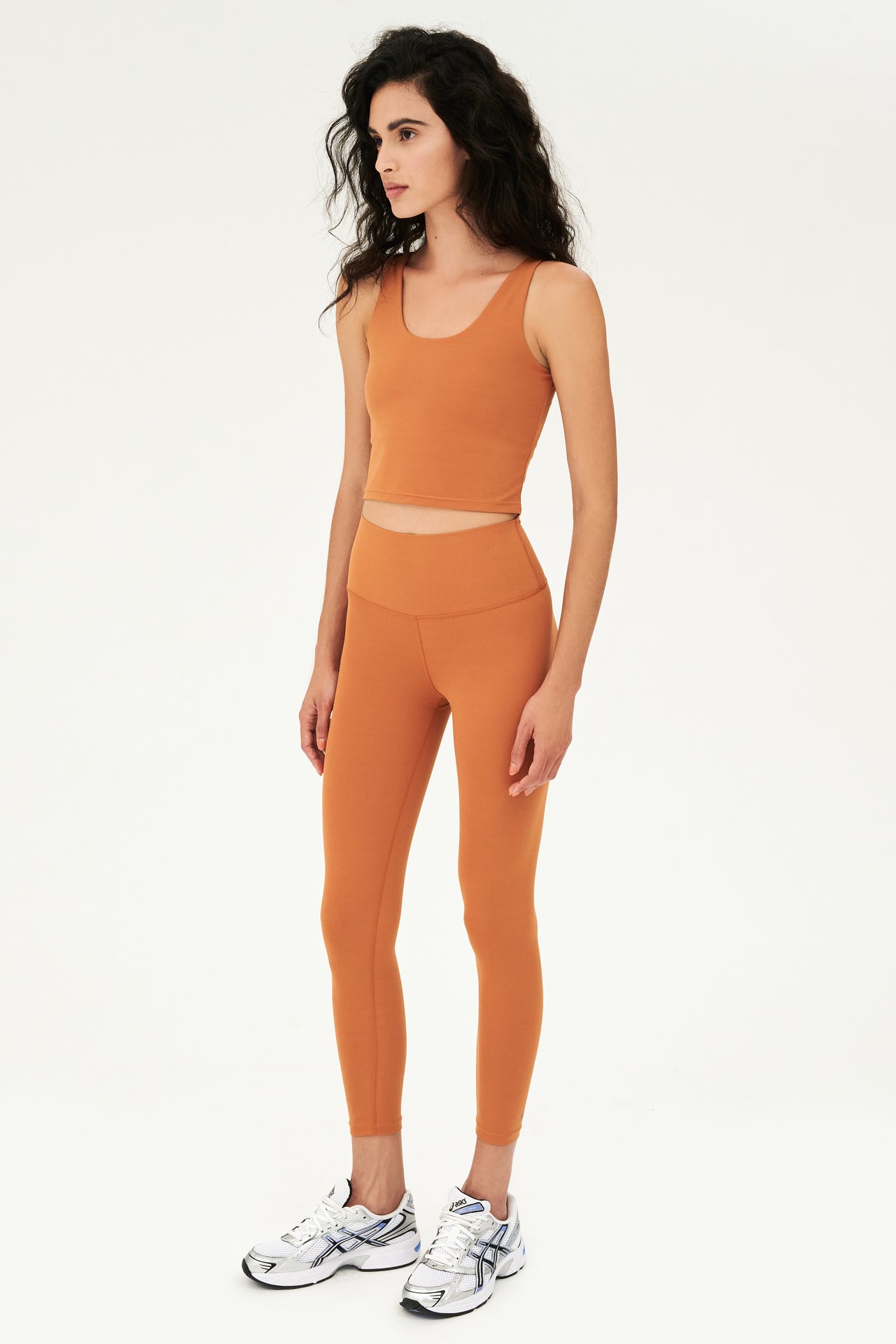 Front full side view of woman with curly dark brown hair wearing a dark orange bra tank with dark orange high waist leggings and white shoes with black stripes