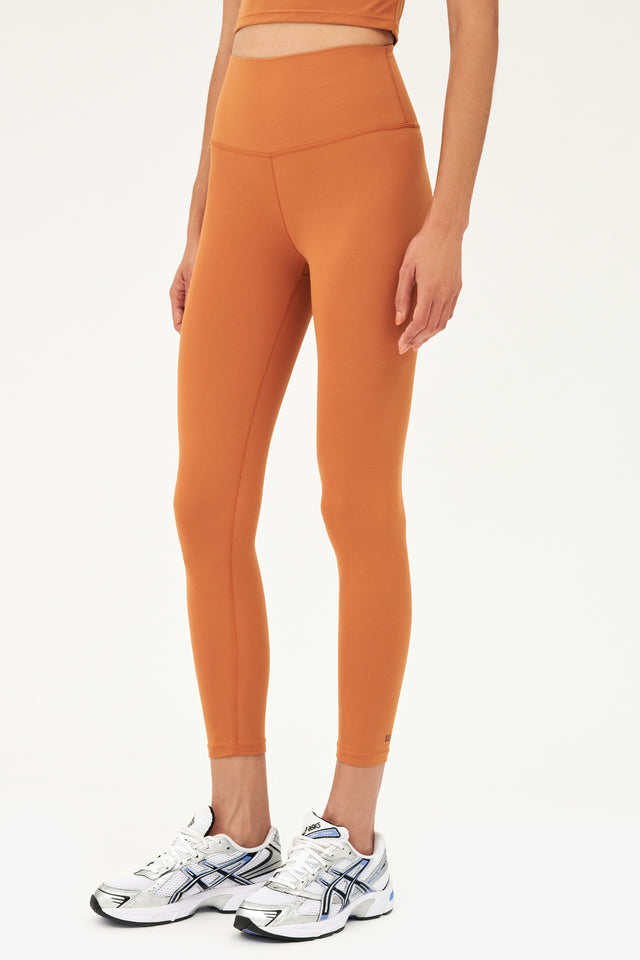 Front full view of model wearing a dark orange bra tank with dark orange high waist leggings and white shoes with black stripes