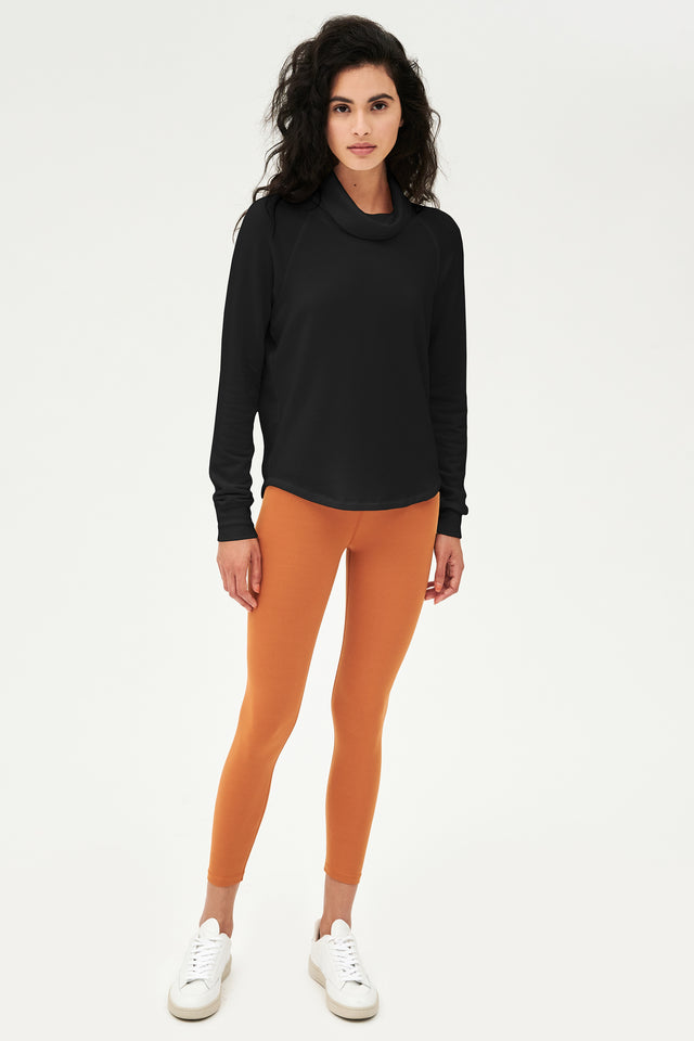 Full front shot of woman with dark wavy hair, wearing black sweatshirt with black cowl neck collar and wearing orange leggings  and white shoes