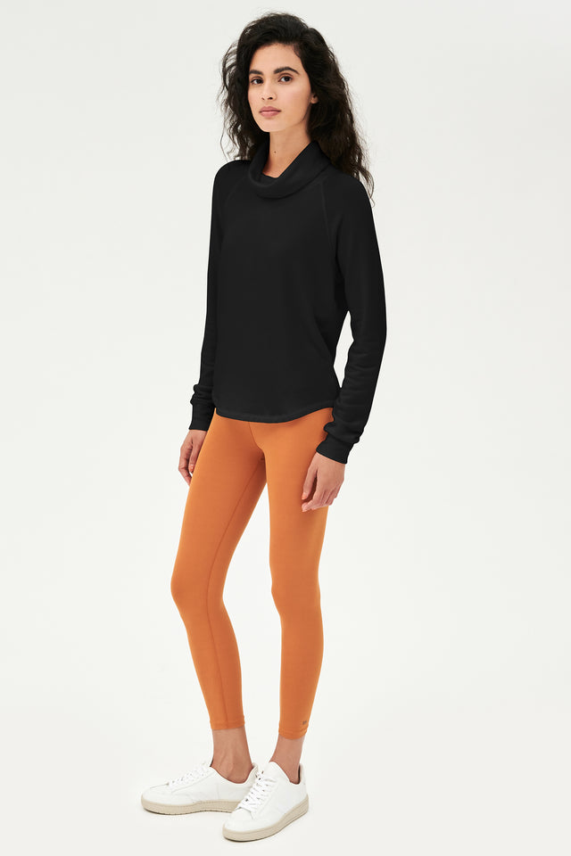 Full side front shot of woman with dark wavy hair, wearing black sweatshirt with black cowl neck collar and wearing orange leggings  and white shoes