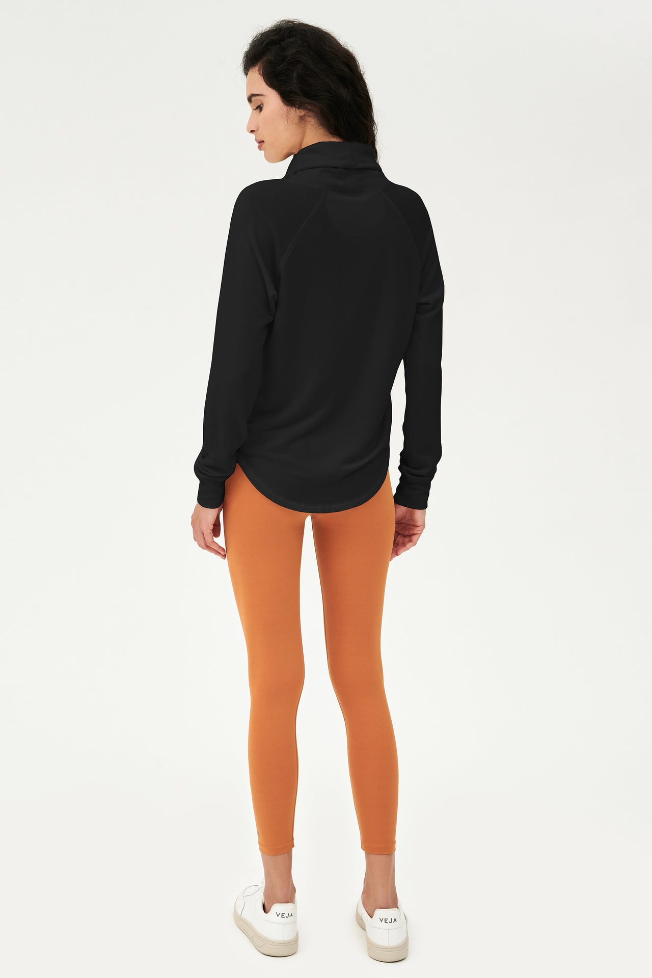 Full back shot of woman with dark wavy hair, wearing black sweatshirt with black cowl neck collar and wearing orange leggings  and white shoes