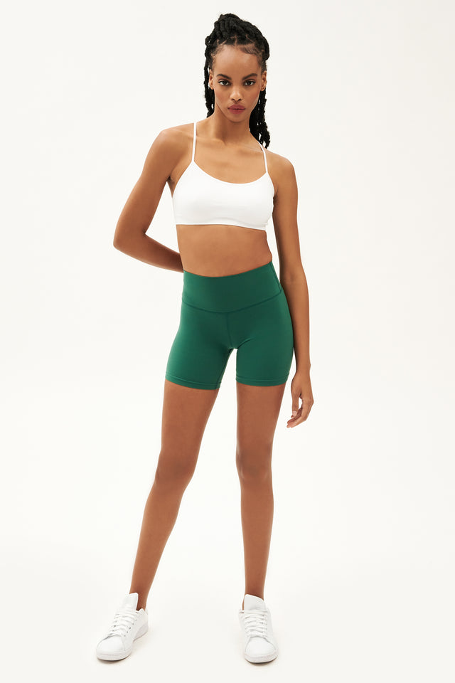 Full front view of girl wearing highwaisted mid thigh green bike shorts with white sports bra and  white shoes
