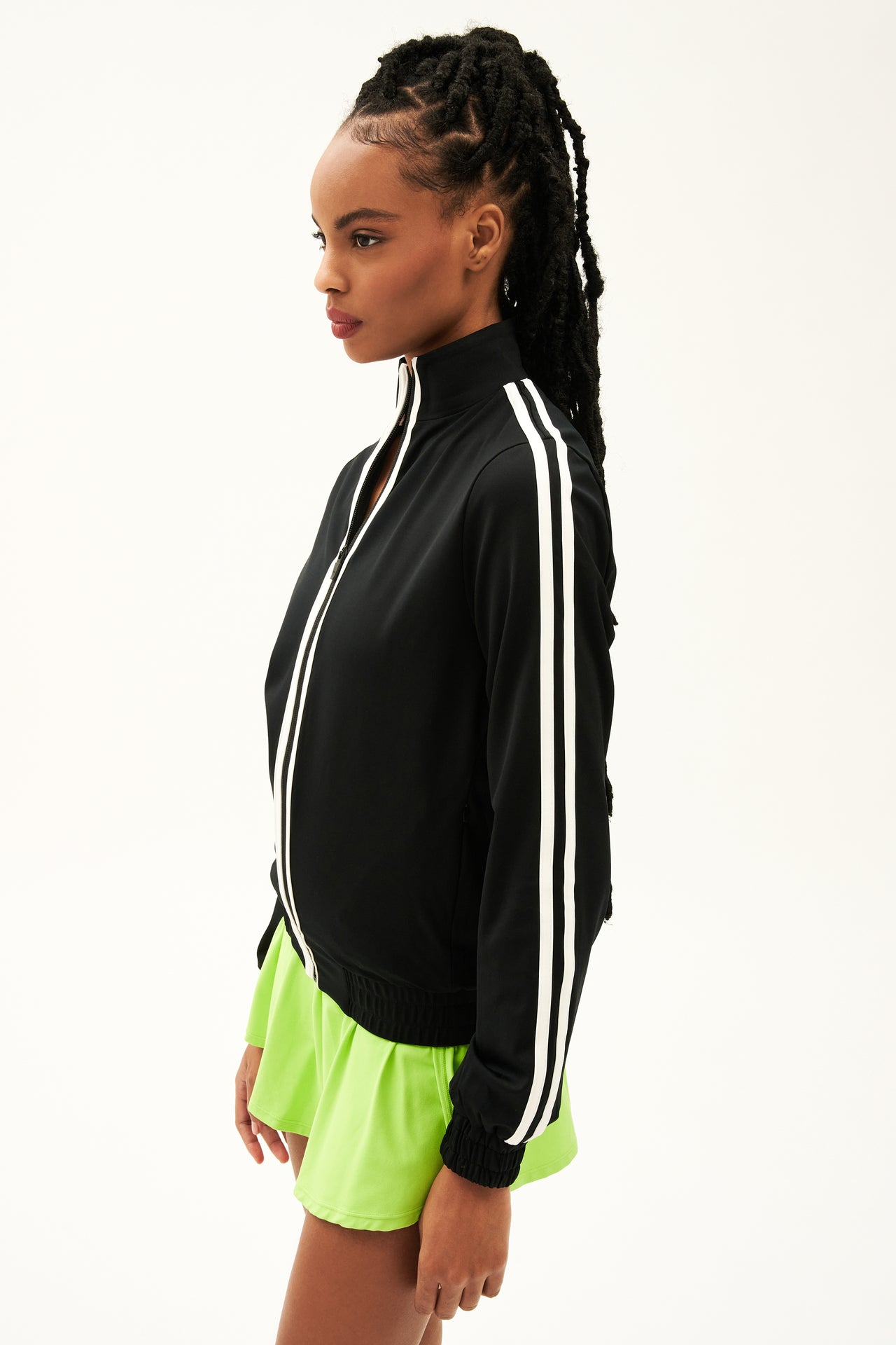 Side view of girl wearing black zip jacket that stops under chin with two white stripes down the side and the front with a  green skirt 