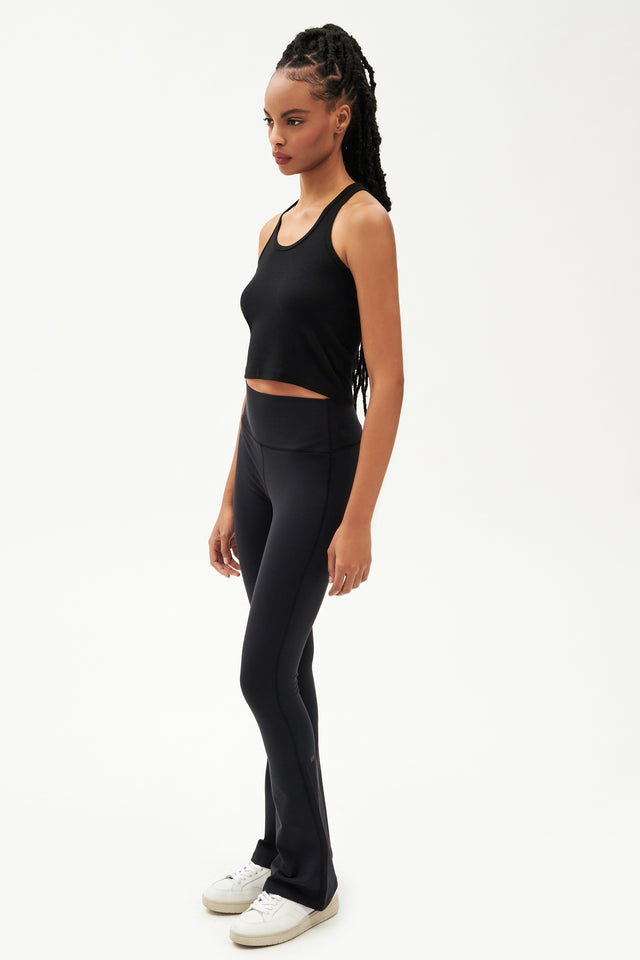 Full side view of girl wearing a ribbed black cropped tank top and black flared leggings with white shoes