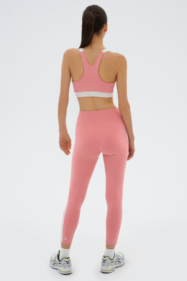 Full back view of girl wearing light pink leggings with a white stripe down the side and inseam with a light pink sports bra with white straps on top of the shoulders and around ribs and multicolored shoes
