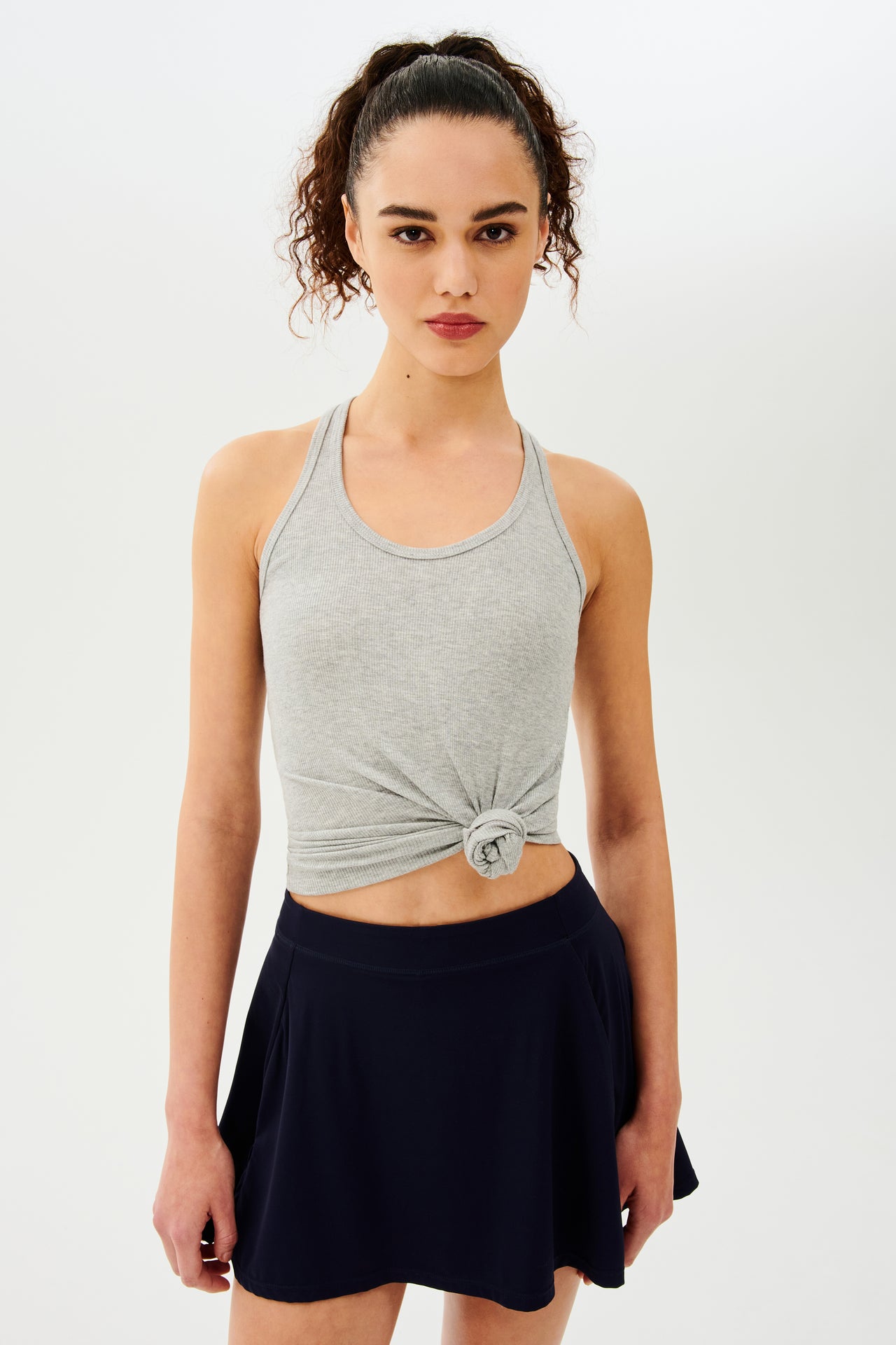 Front view of girl wearing a light grey tank top tied in a knot at the waist and black skirt 