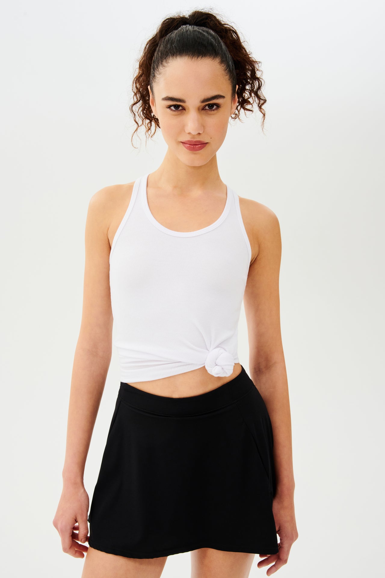 Front view woman with dark curly hair in a ponytail wearing a white tank top with a tied knot and black skort 