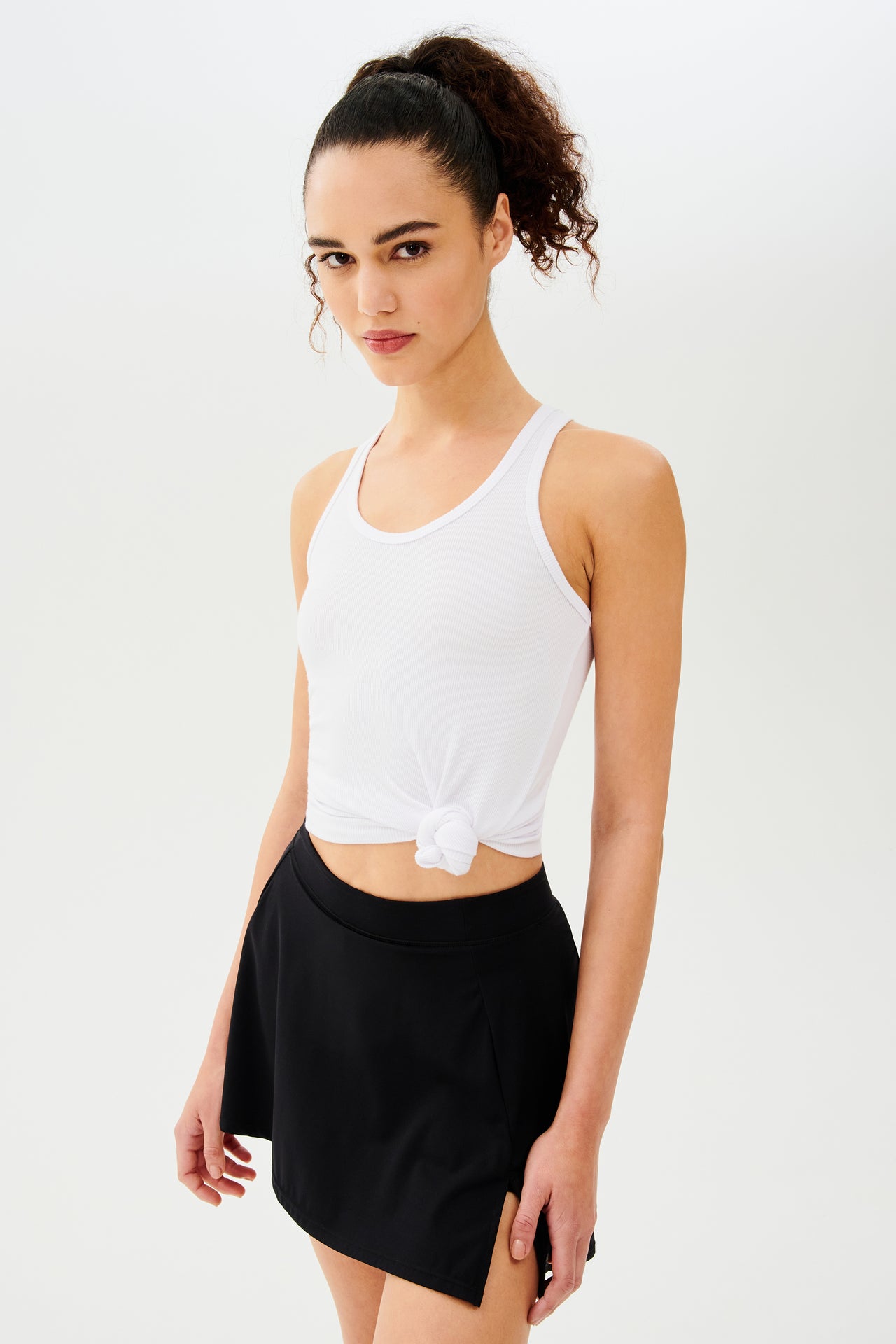 Front side view woman with dark curly hair in a ponytail wearing a white tank top with a tied knot and black skort 