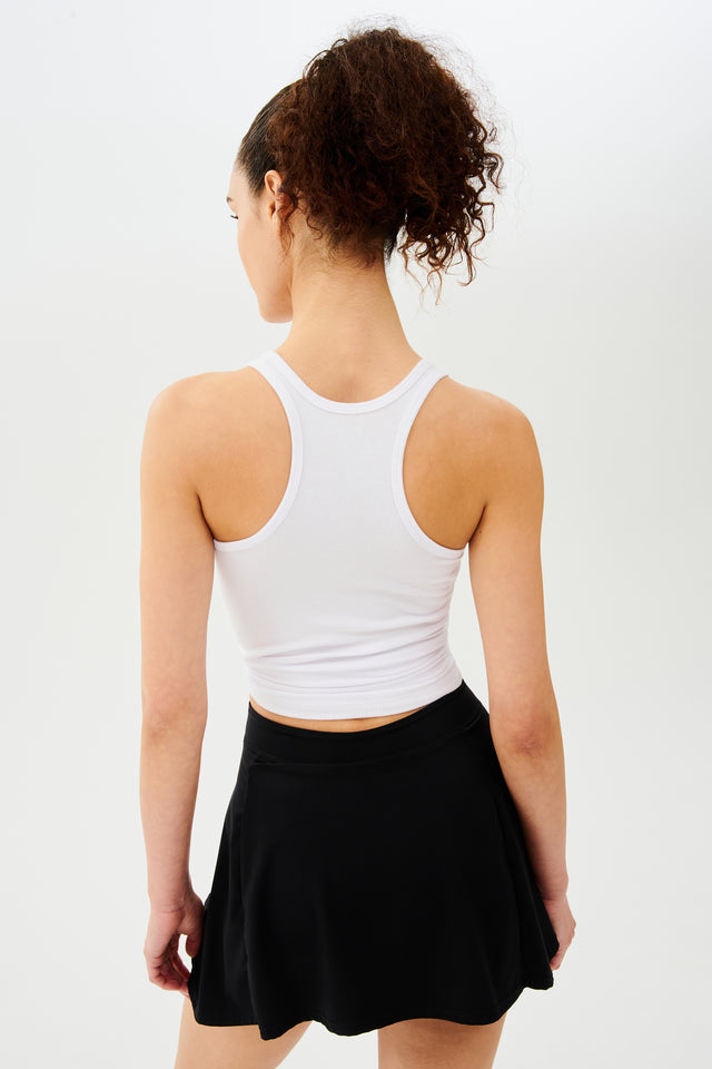 Back view woman with dark curly hair in a ponytail wearing a white tank top and black skort