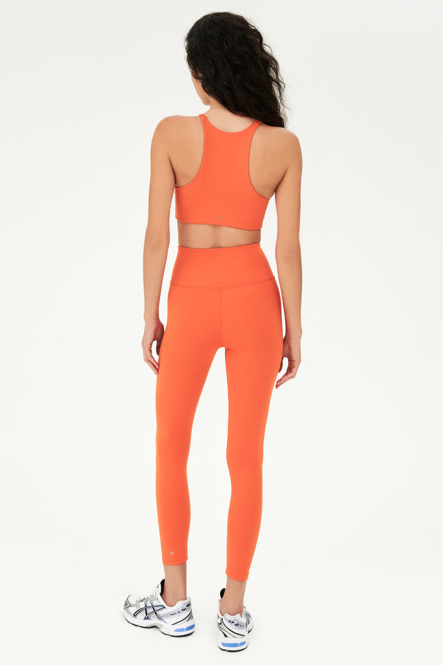 Full back view of girl wearing orange sports bra that stops at collarbone and orange leggings with white shoes