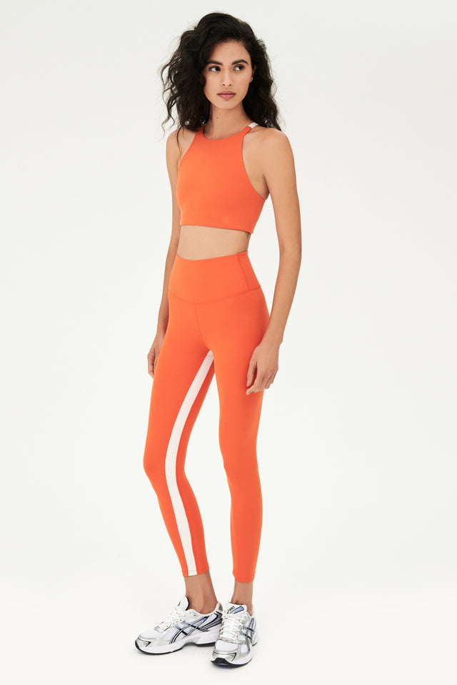 Full side view of girl wearing orange sports bra that stops at collarbone and orange leggings with white shoes