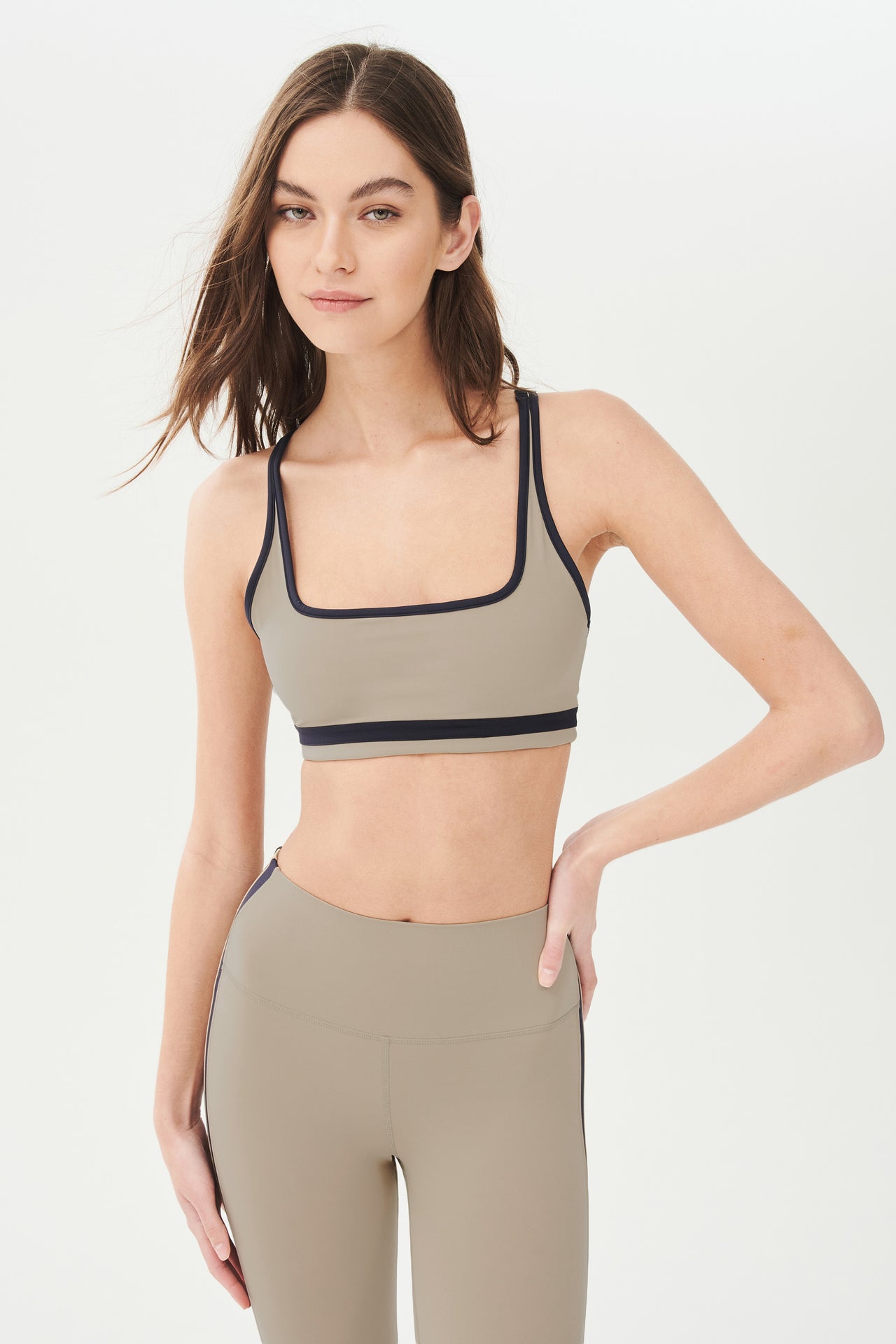 Front view of girl wearing dark light brown sports bra with black colored stripe along the ribs and black hem around the arms and neck with dark light brown leggings with two black stripes down the side 