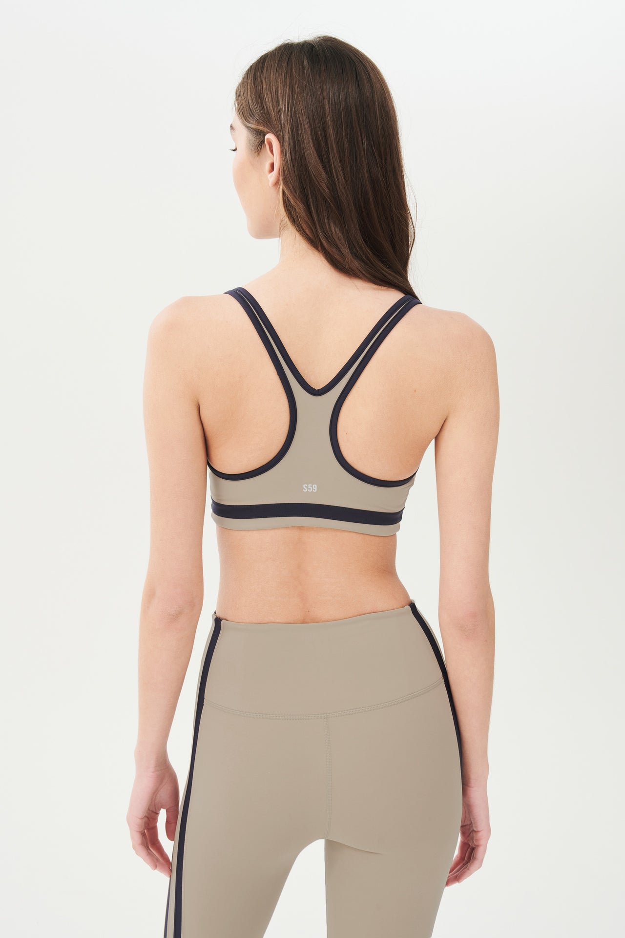 Back view of girl wearing dark light brown sports bra with black colored stripe along the ribs and black hem around the arms and neck with dark light brown leggings with two black stripes down the side 