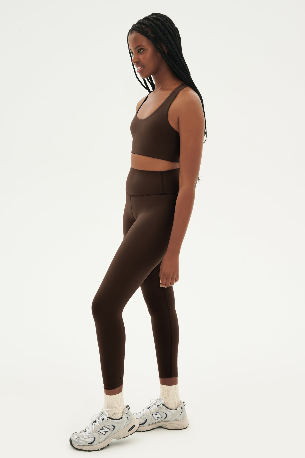 Full front side view of woman with black braids wearing dark brown bra and dark brown leggings paired with white shoes with black stripes 